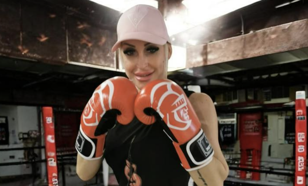 Boxer wearing a pink cap and black top, gloves up, posing in a gym boxing ring.