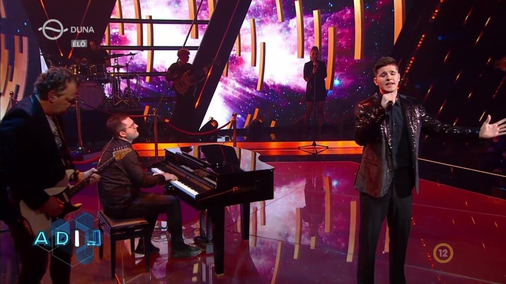 Male singer in a sequined jacket performs on a glittery TV stage with a pianist and guitarist, spacey background.