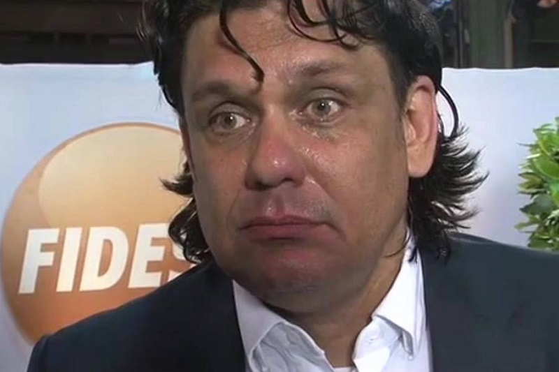 Close-up of a man in a dark suit and white shirt, sweating and with a distressed expression during an interview, with a blurred orange logo in the background.
