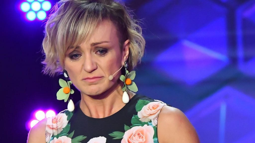 A woman performer on stage with a floral sleeveless dress and a headset mic, looking down with a somber expression.